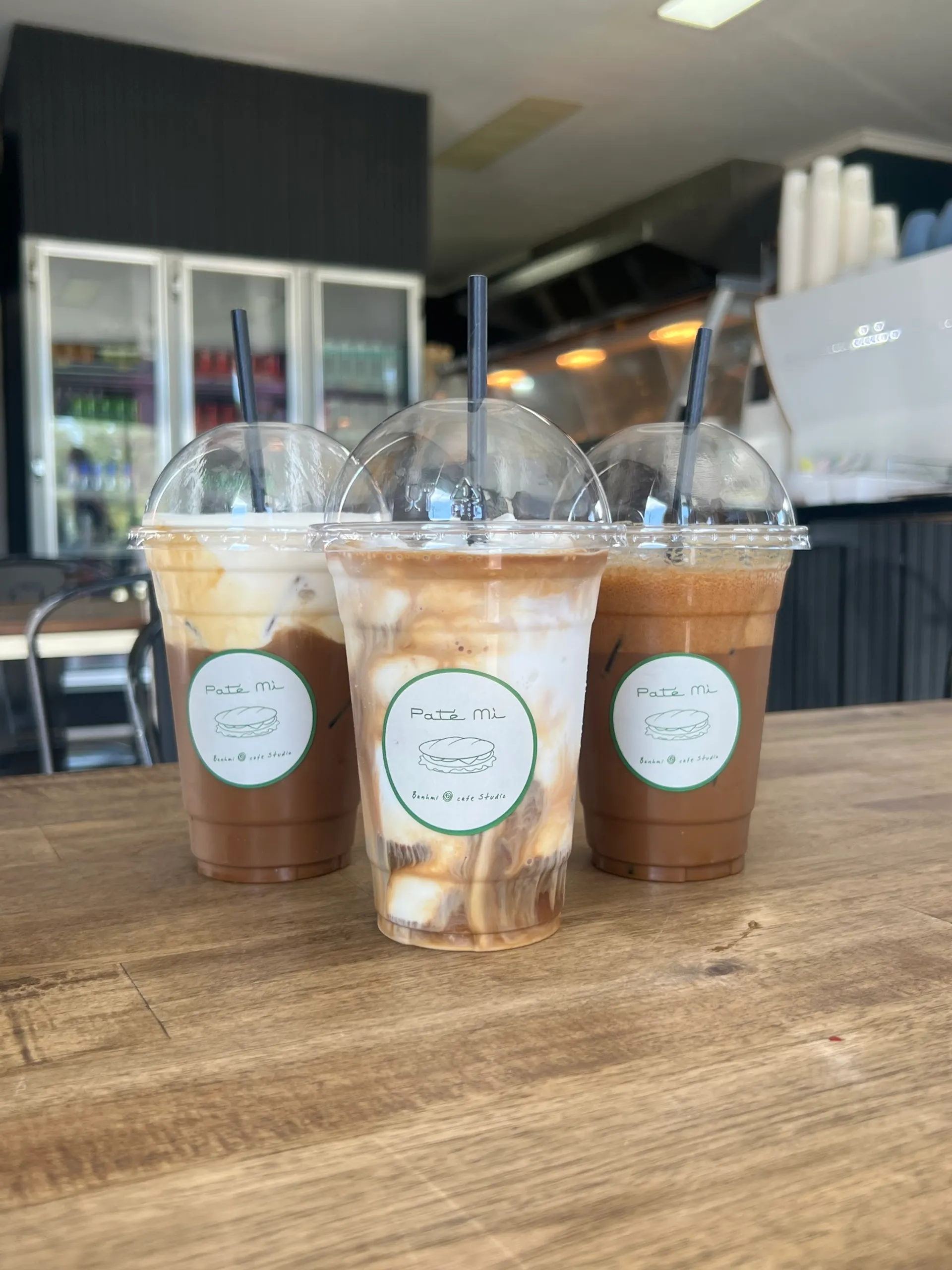 Three branded Pate Mi iced coffees on a wood table with cafe counter blurred behind