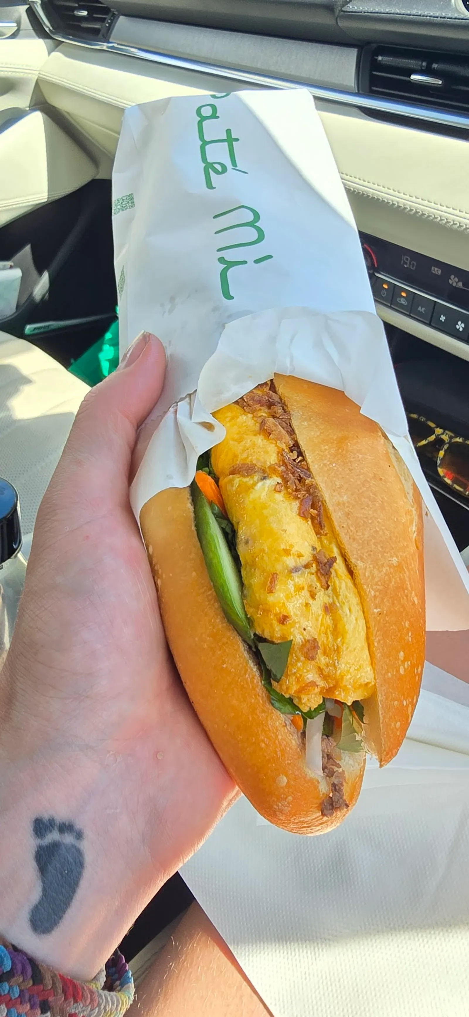 Hand holding an omelette banh mi in a branded Pate Mi paper sleeve inside a car